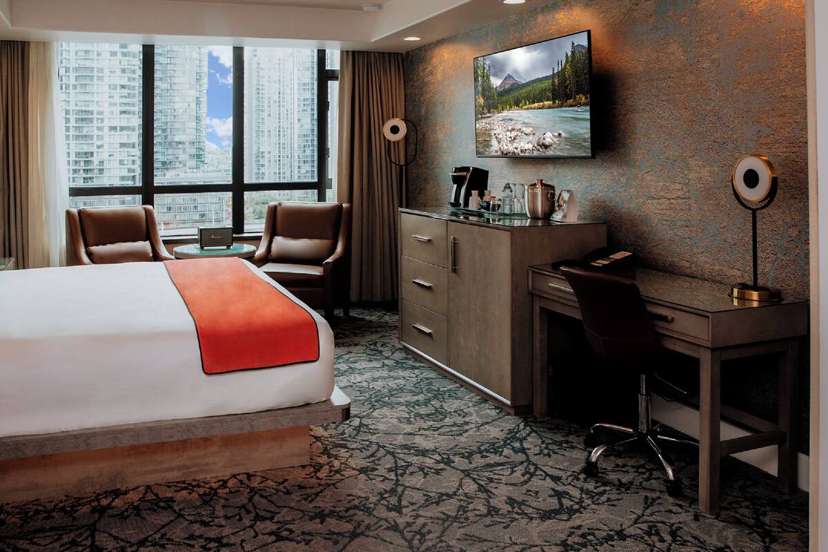 A hotel room with a large bed, a desk, a chair, a television, and a view of a cityscape through the window.