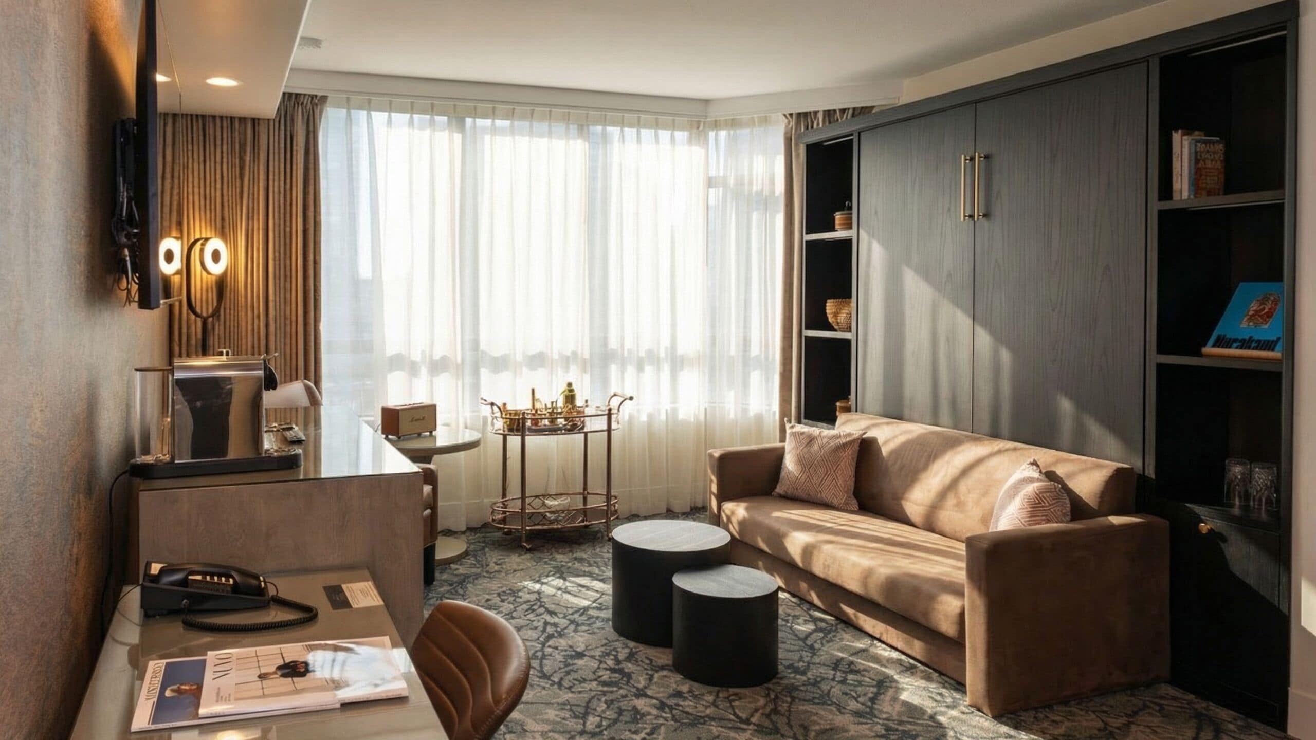 A modern hotel room with a leather sofa, a desk with a telephone, a small round table, and a bookshelf.