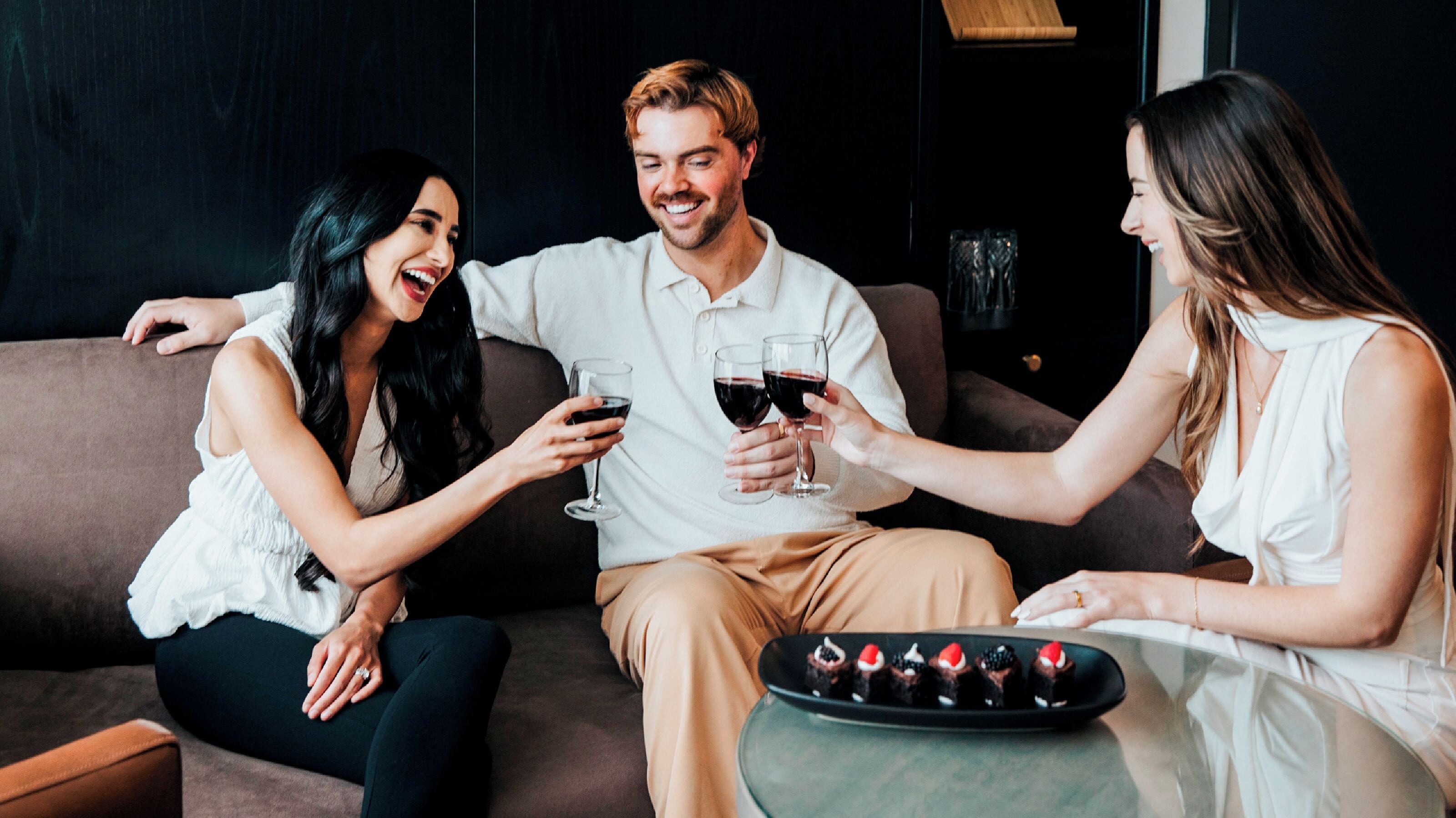 Three people sitting on a couch, holding glasses of red wine, with a tray of desserts in front of them.