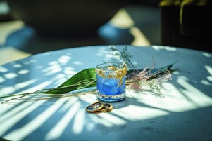 A glass of blue liquid with gold flakes on a table with a leaf and a decorative item.
