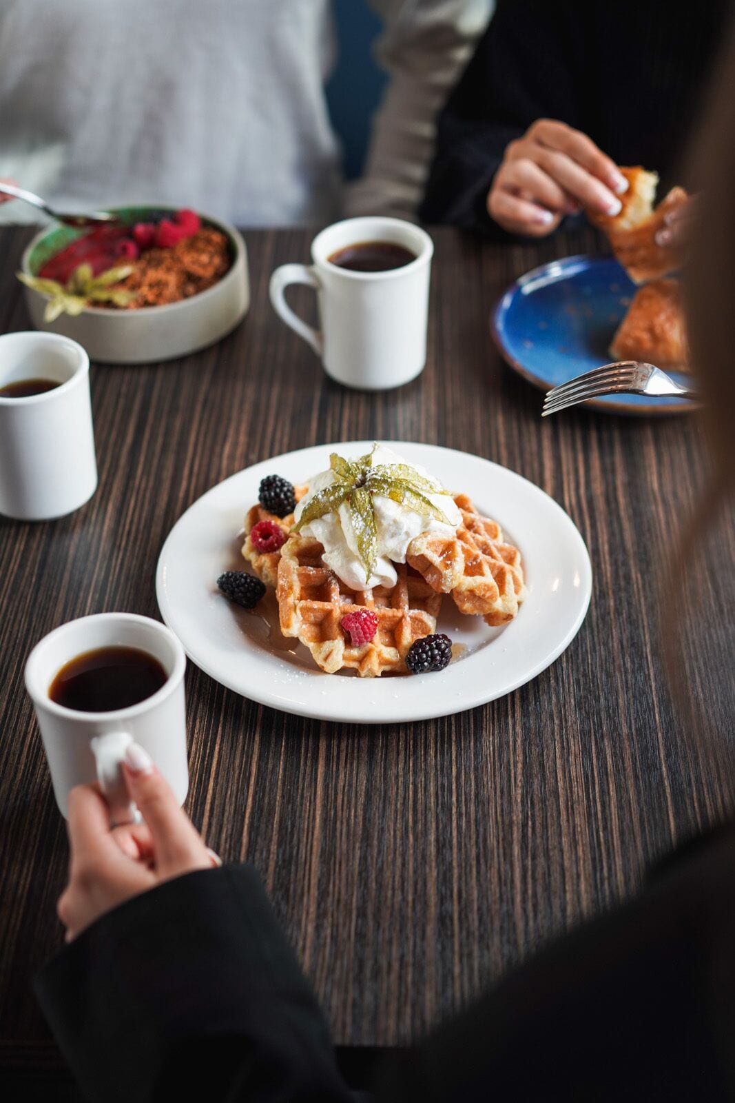 Une assiette de gaufres garnies de crème fouettée et de fruits rouges, une tasse de café et un saladier rempli de salade, le tout posé sur une table en bois.