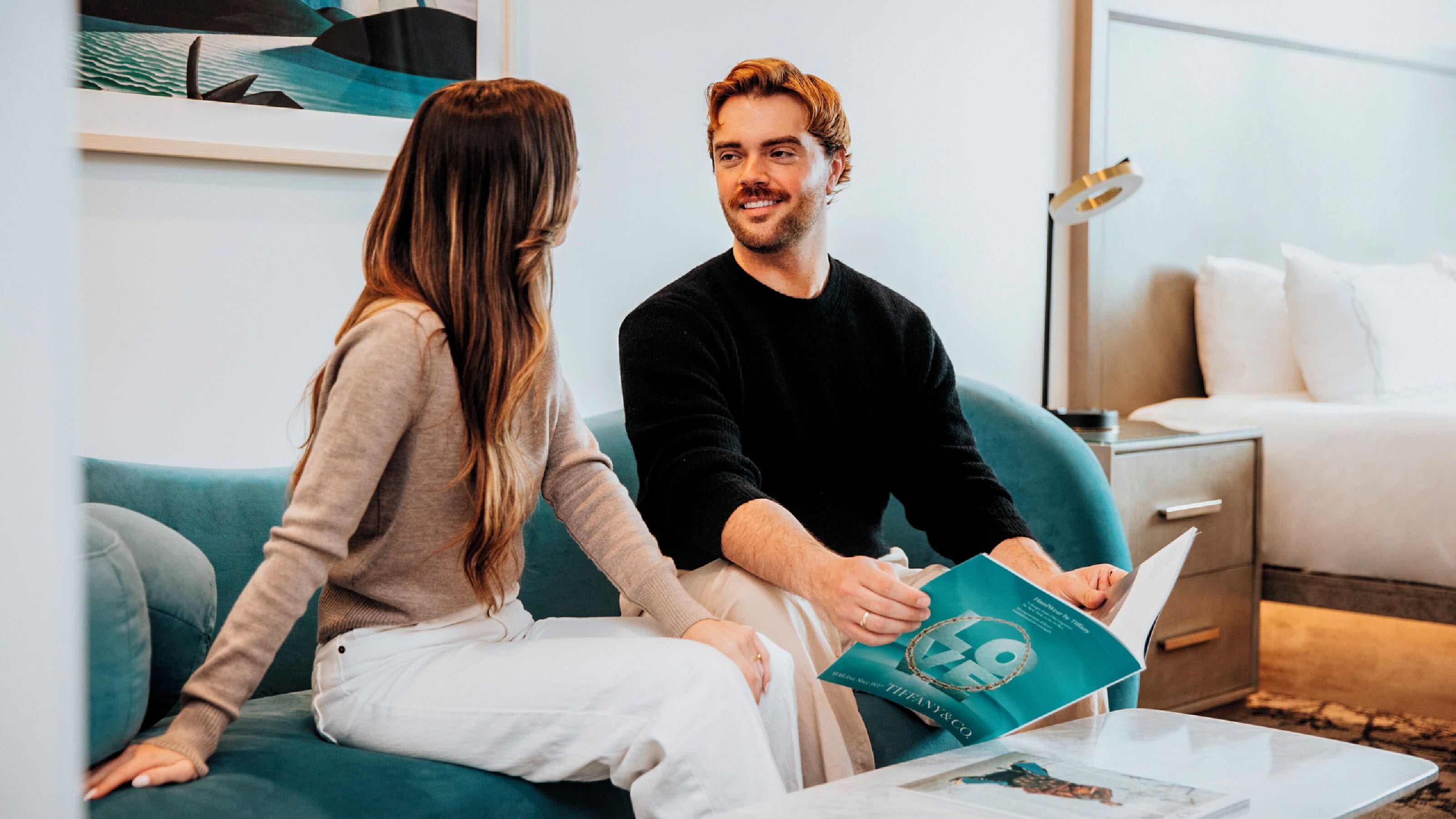 A couple sitting on a teal sofa in a modern living room with a minimalist design, a coffee table, and a bed in the background.