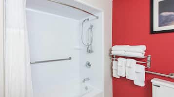 Baño con tina blanca, ducha y sanitario. Hay un perchero con toallas dobladas.
