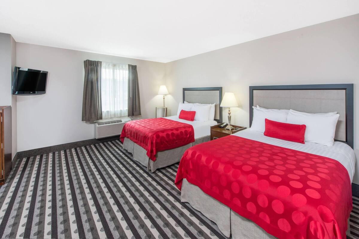 A hotel room with two beds, each with a red polka dot bedspread, a flat-screen TV mounted on the wall, and a window with curtains.
