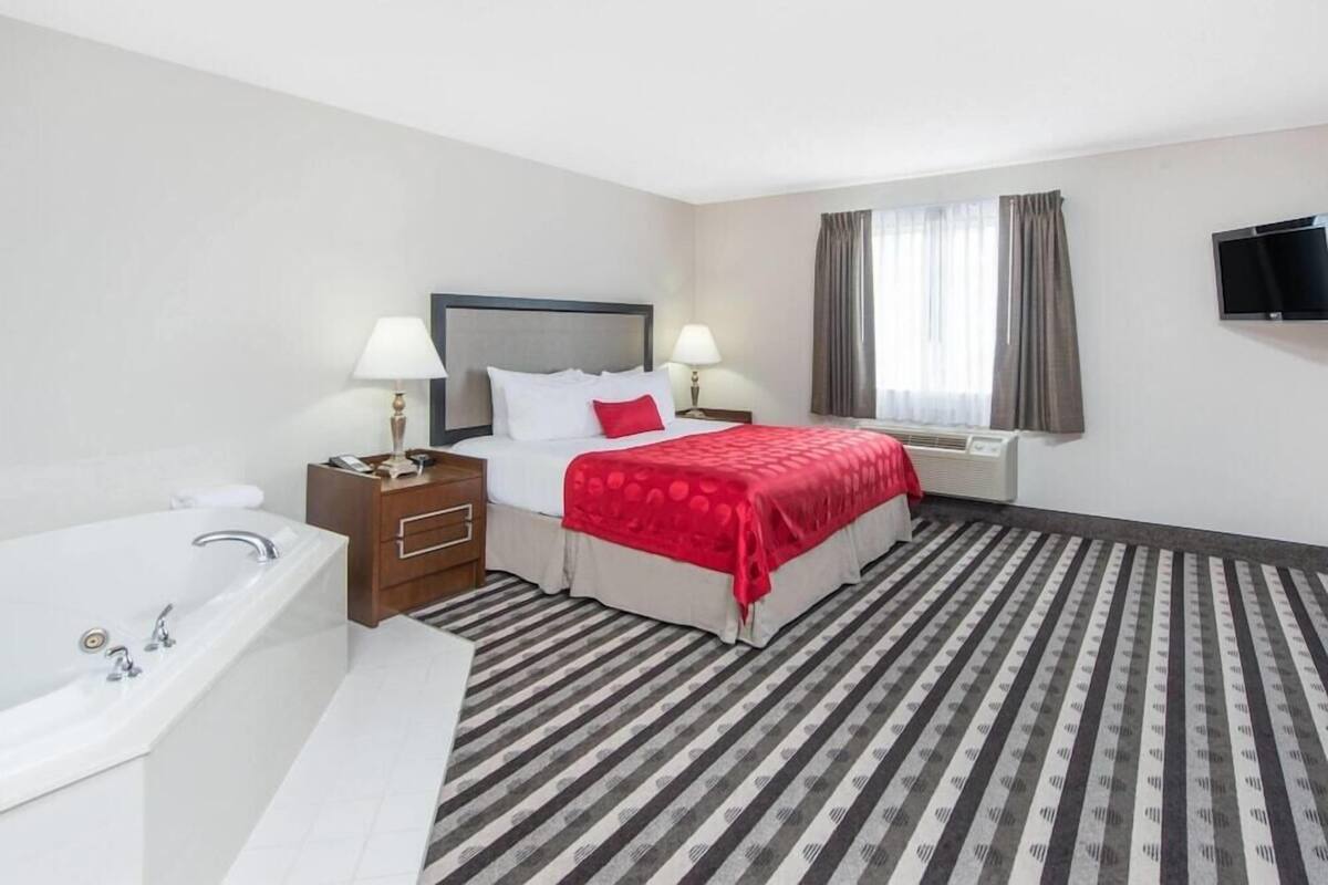 A hotel room with a bed, two bedside tables, a TV, a window with curtains, a bathroom with a bathtub, and a striped carpet.