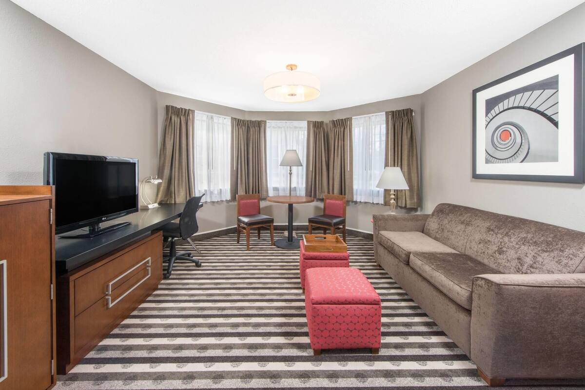 A hotel room with a flat-screen TV, a wooden desk, a sofa, a patterned rug, and a painting of a spiral staircase.