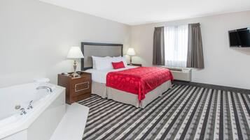 Quarto de hotel com uma cama, duas mesas de cabeceira com luminárias, uma TV fixada na parede, uma janela com cortinas, um banheiro com banheira e pia, e um tapete listrado.