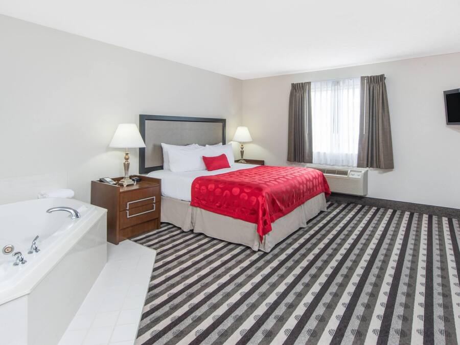 Quarto de hotel com uma cama, duas mesas de cabeceira com luminárias, uma TV fixada na parede, uma janela com cortinas, um banheiro com banheira e pia, e um tapete listrado.
