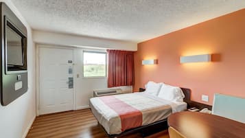 Une chambre d’hôtel comprenant un lit, un bureau, une chaise, une fenêtre avec des rideaux, un radiateur fixé au mur et une porte avec une pancarte.