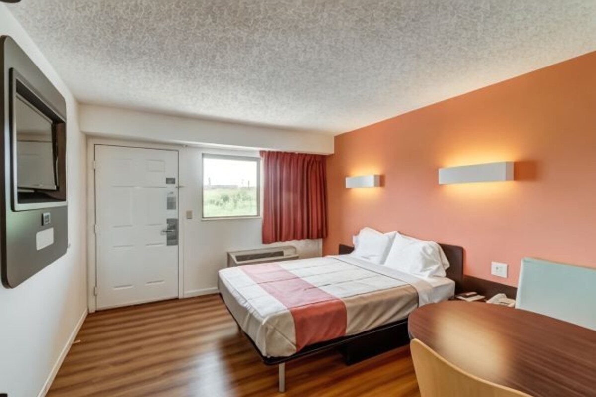 Habitación de hotel con cama, escritorio, silla, ventana con cortinas, aire acondicionado en la pared y una puerta.