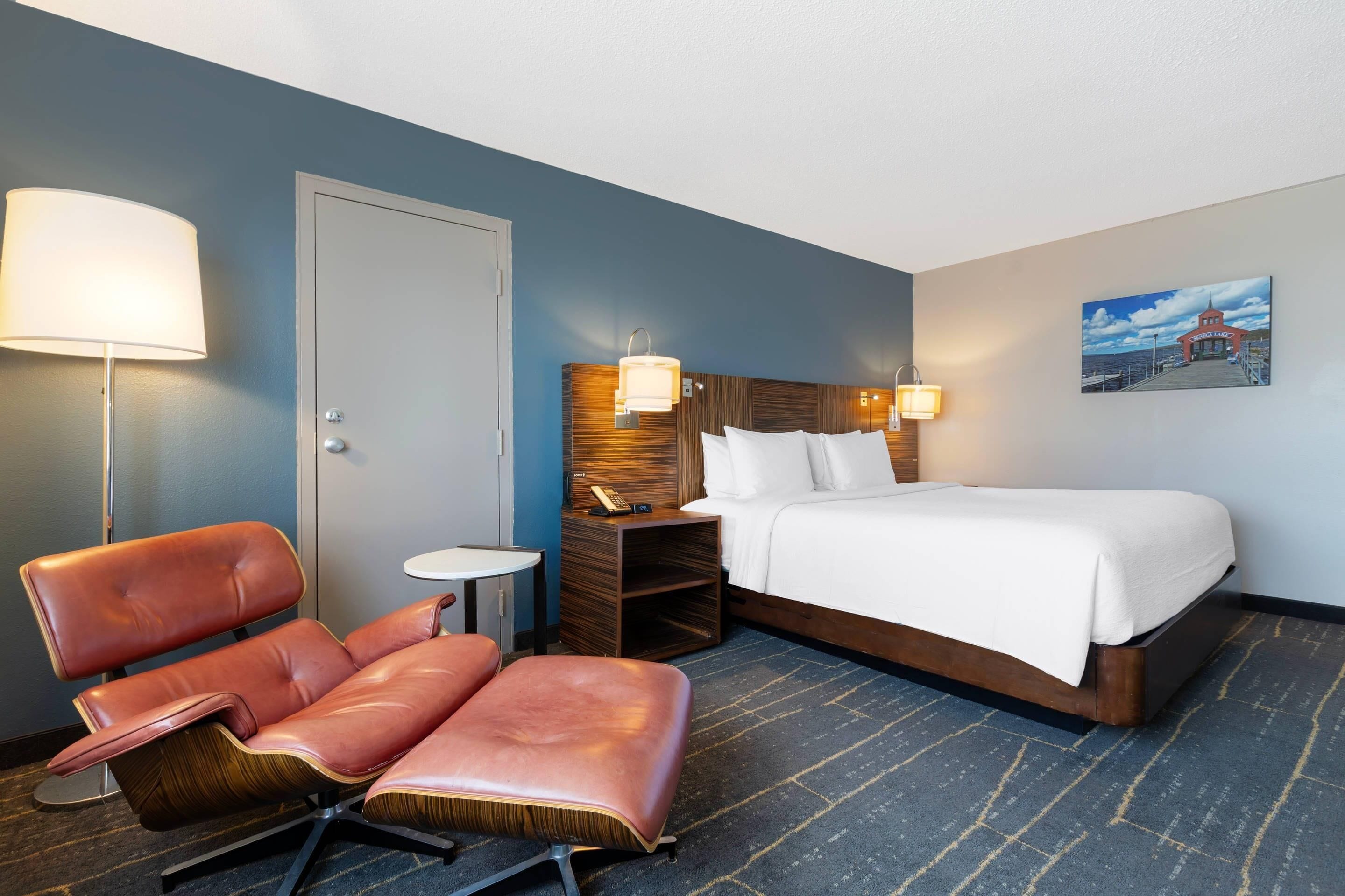 A hotel room with a bed, a desk, a chair, a lamp, and a painting on the wall.
