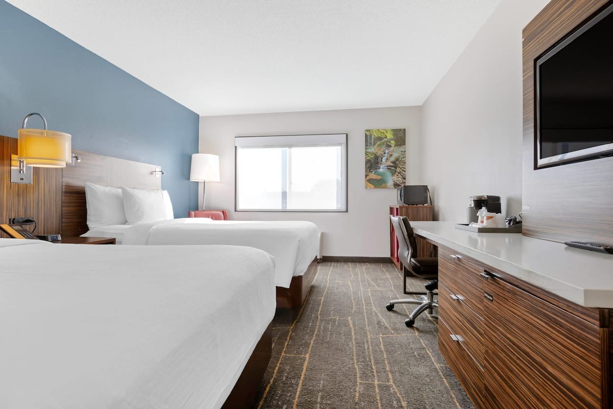 A hotel room with two beds, a desk with a flat-screen TV, a desk lamp, a coffee maker, and a large window.