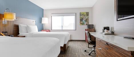A hotel room with two beds, a desk with a flat-screen TV, a desk lamp, a coffee maker, and a large window.