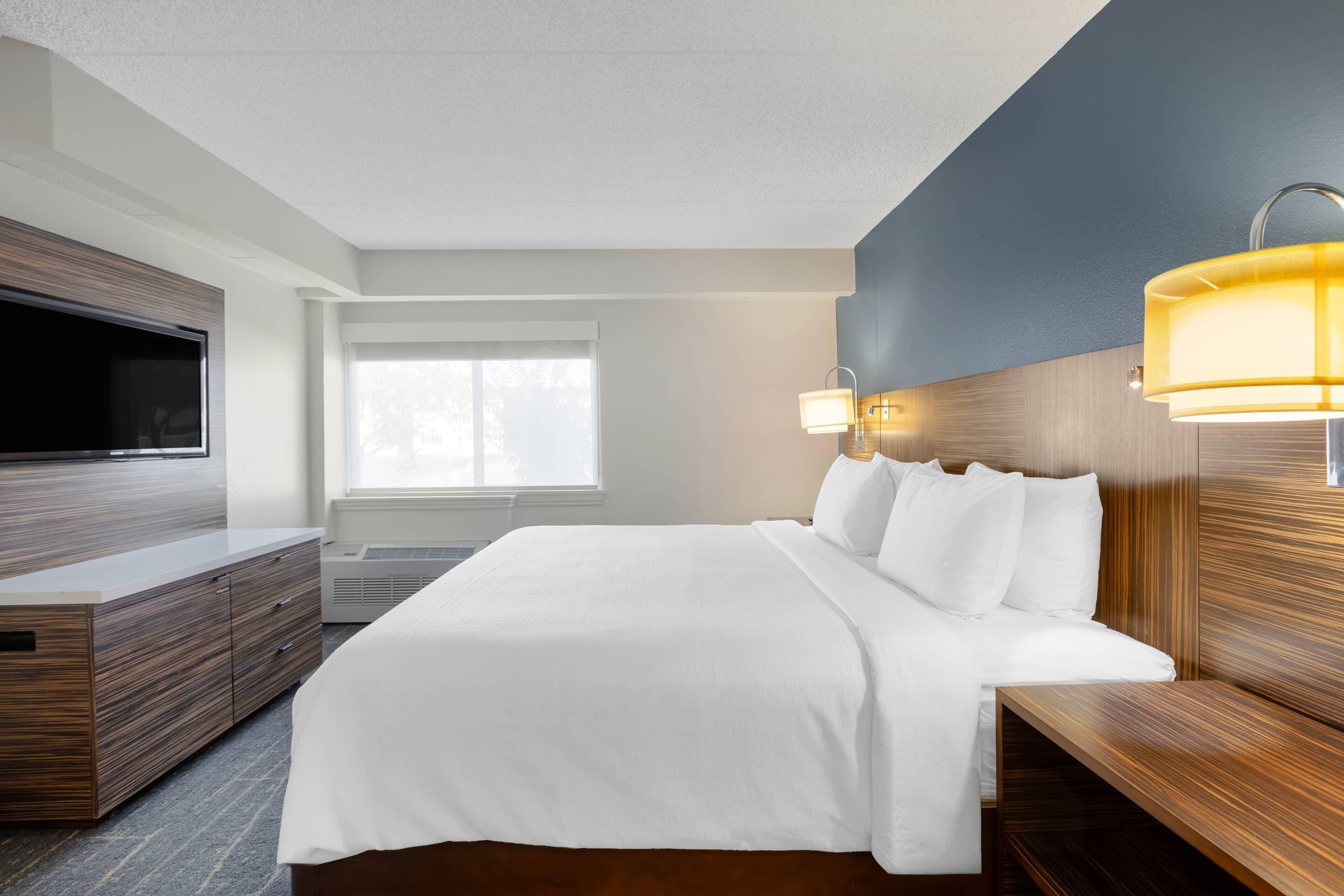 A modern hotel room with a large bed, wooden headboard, a flat-screen TV, and a window providing natural light.