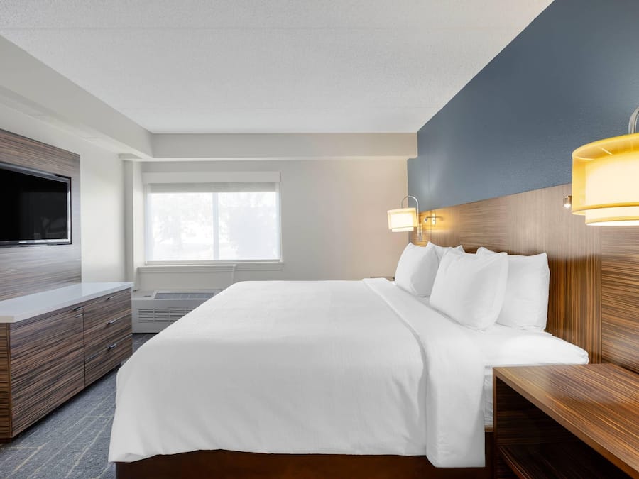 A modern hotel room with a large bed, wooden headboard, a flat-screen TV, and a window providing natural light.