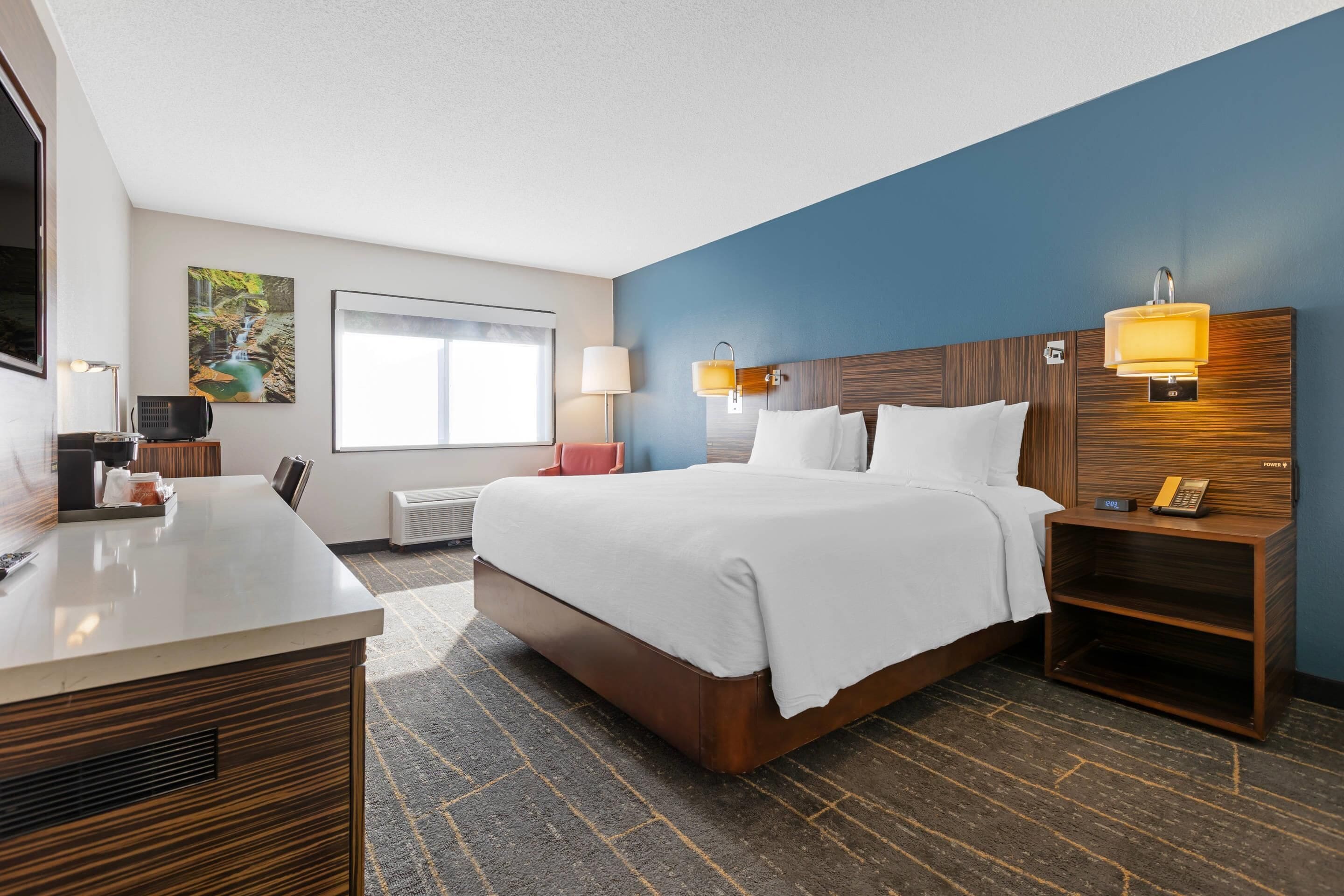 A hotel room with a bed, bedside tables, a desk, a chair, a window, and artwork on the wall.