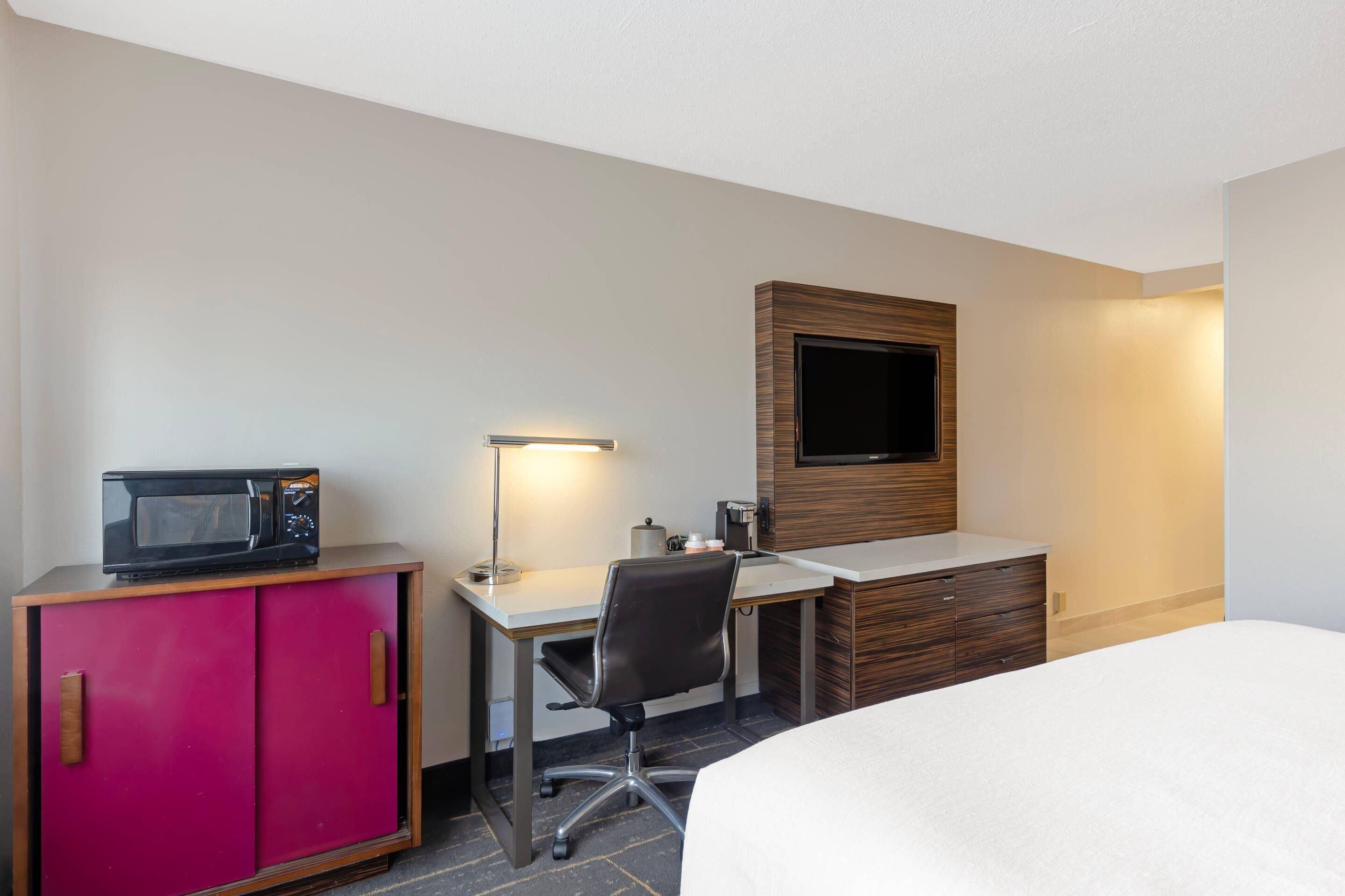 A hotel room with a bed, a desk with a computer, a microwave, a TV, and a minibar.
