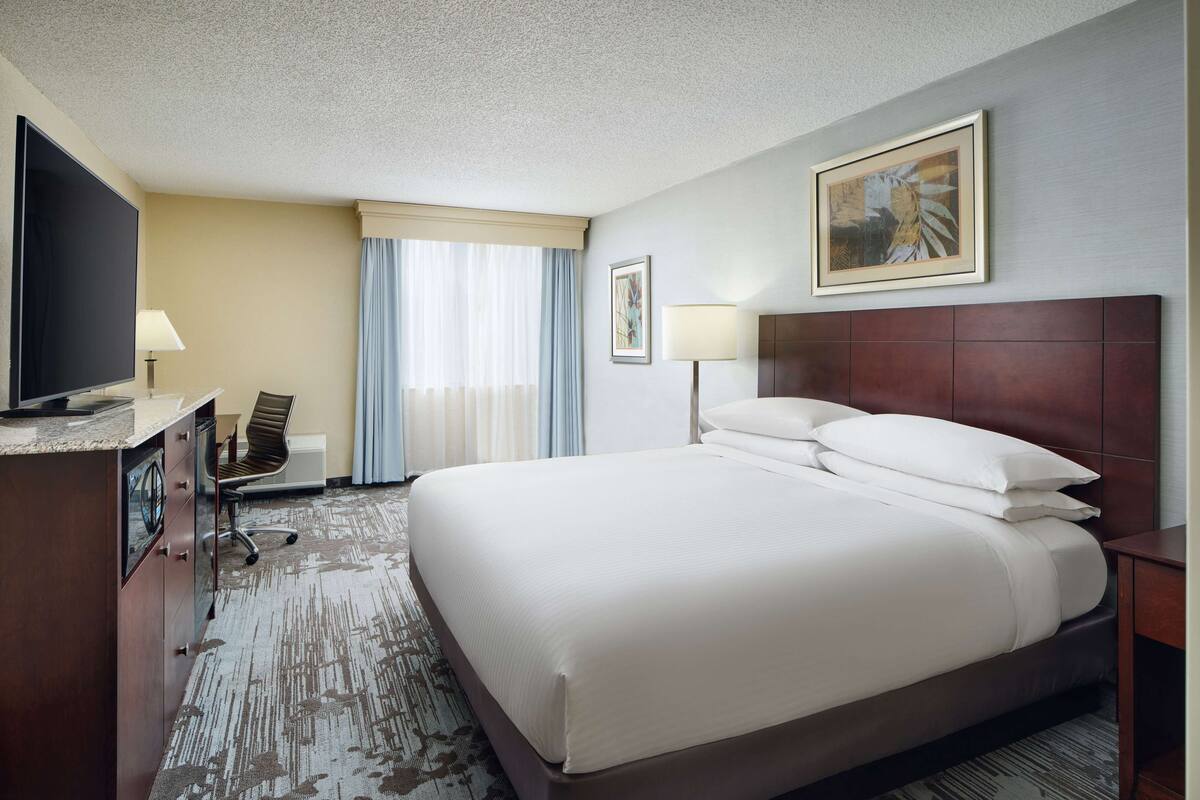 A hotel room with a large bed, a desk with a flat-screen TV, a chair, a desk lamp, and a painting on the wall.
