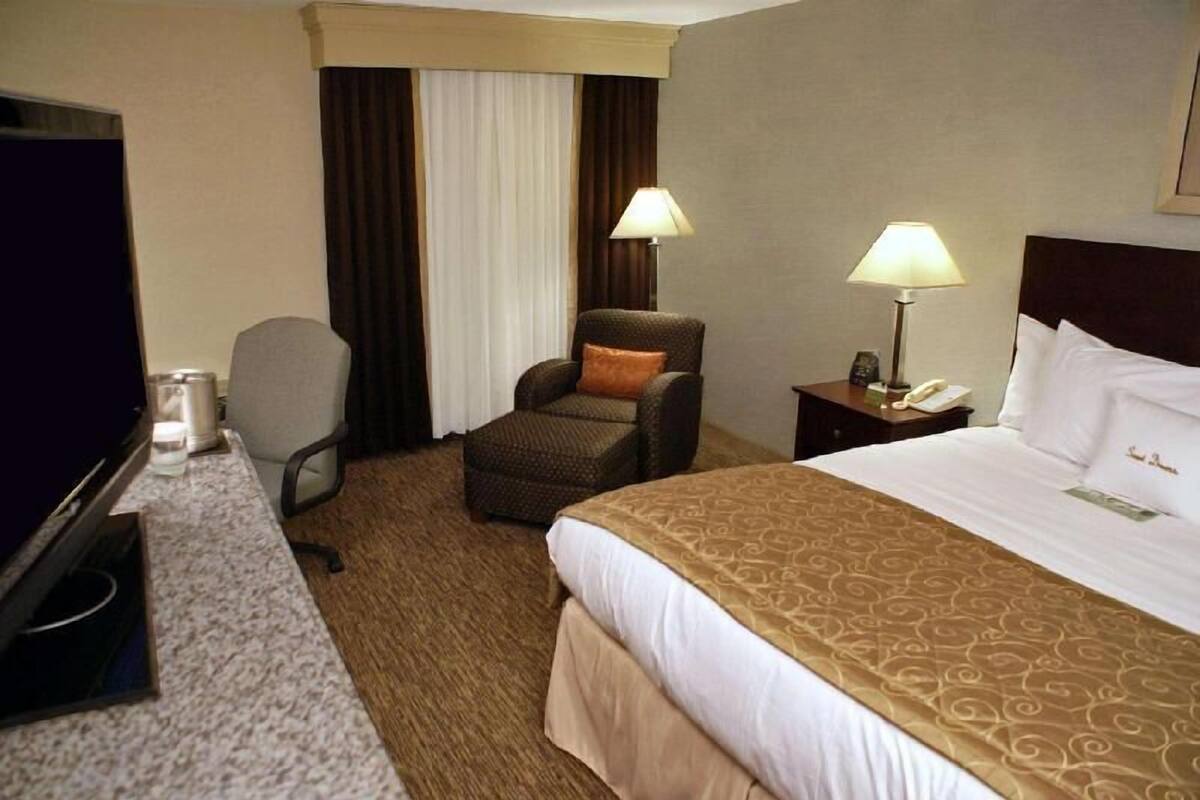 A hotel room with a bed, a chair, a desk with a computer, a lamp, and a nightstand with a phone.