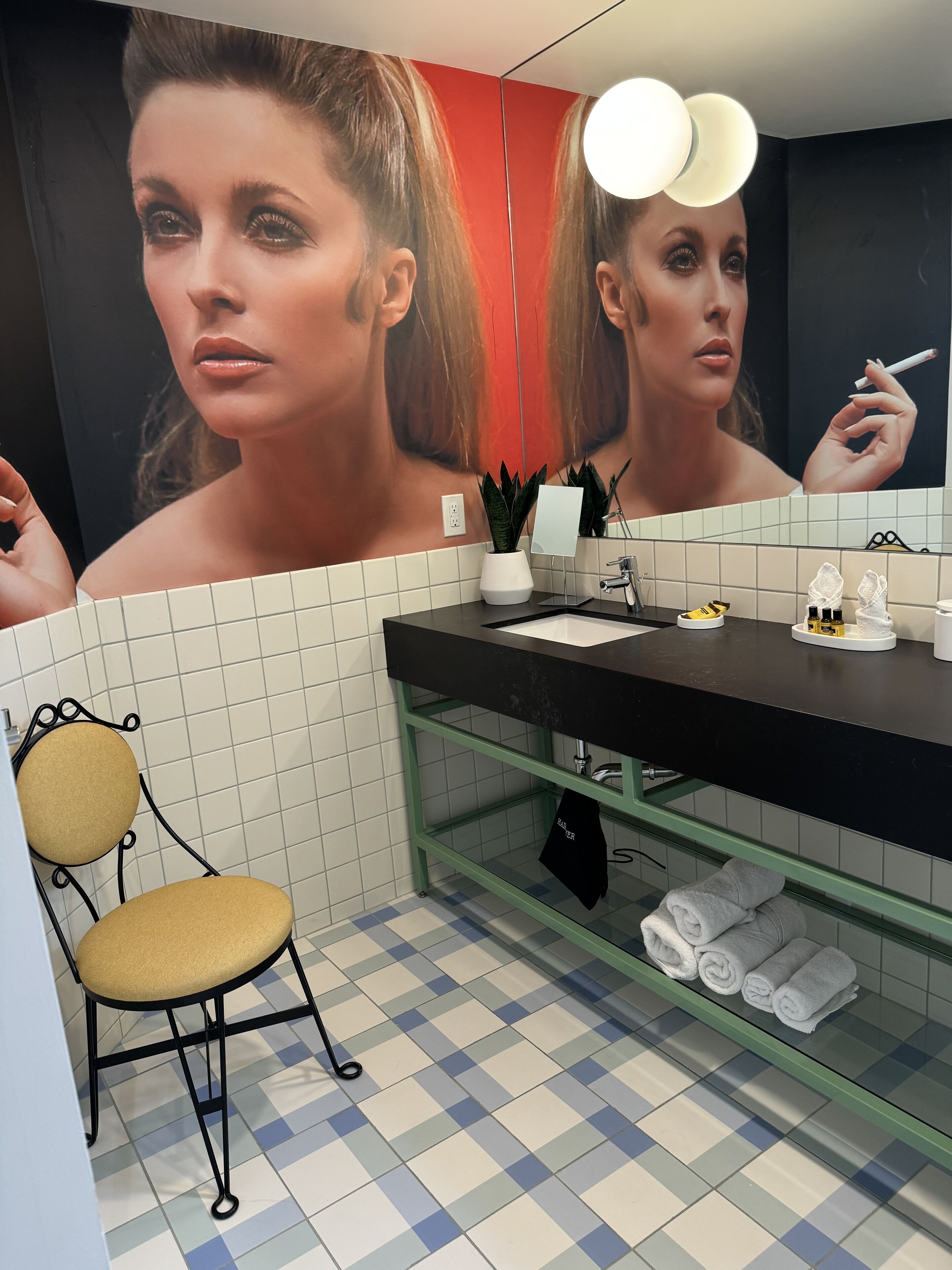 A bathroom with a checkered floor, a black sink, a mirror, a towel rack, and a chair.