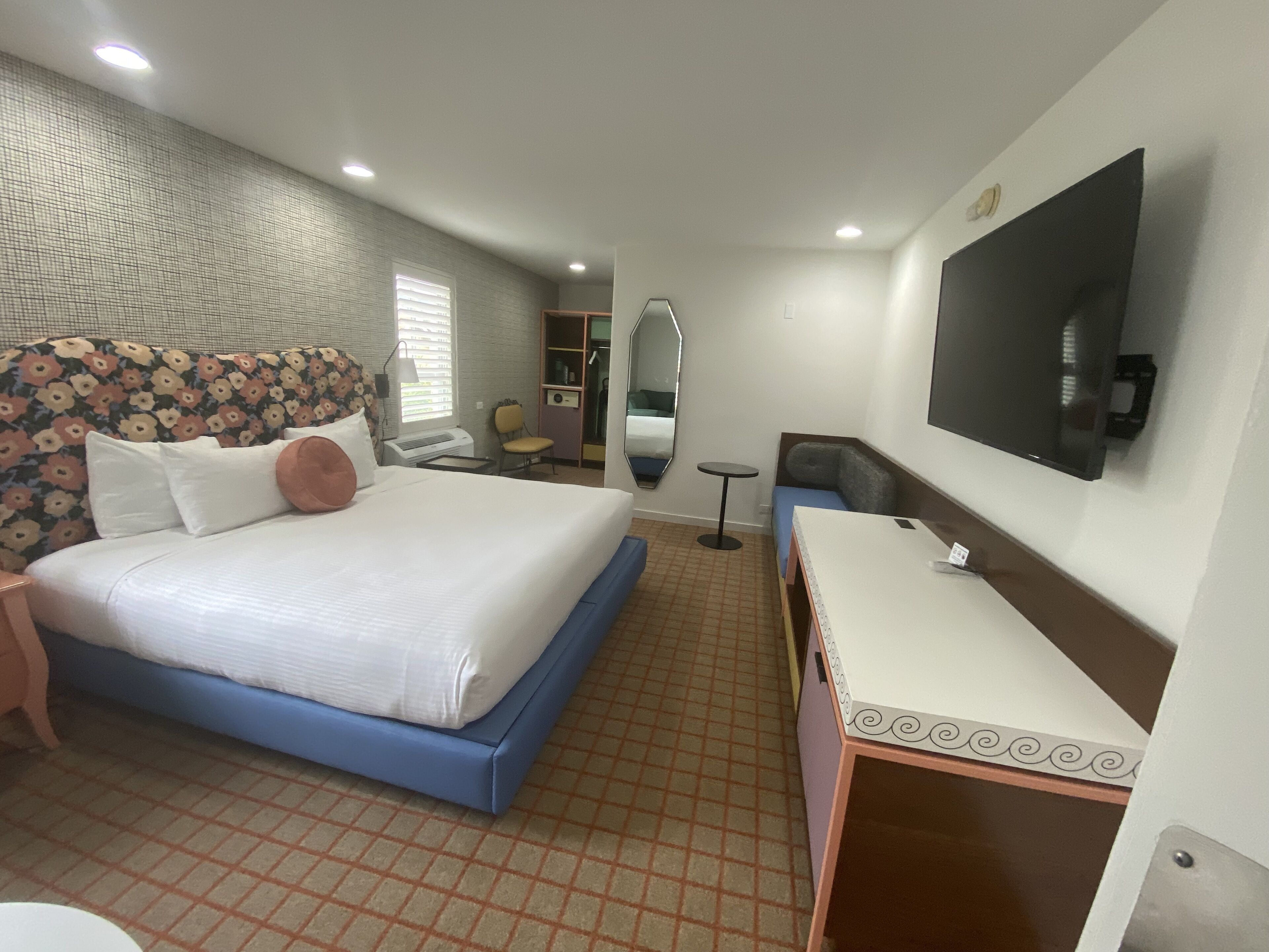 Una habitación de hotel con cama, mesita de noche, espejo, televisor y una pequeña mesa redonda.