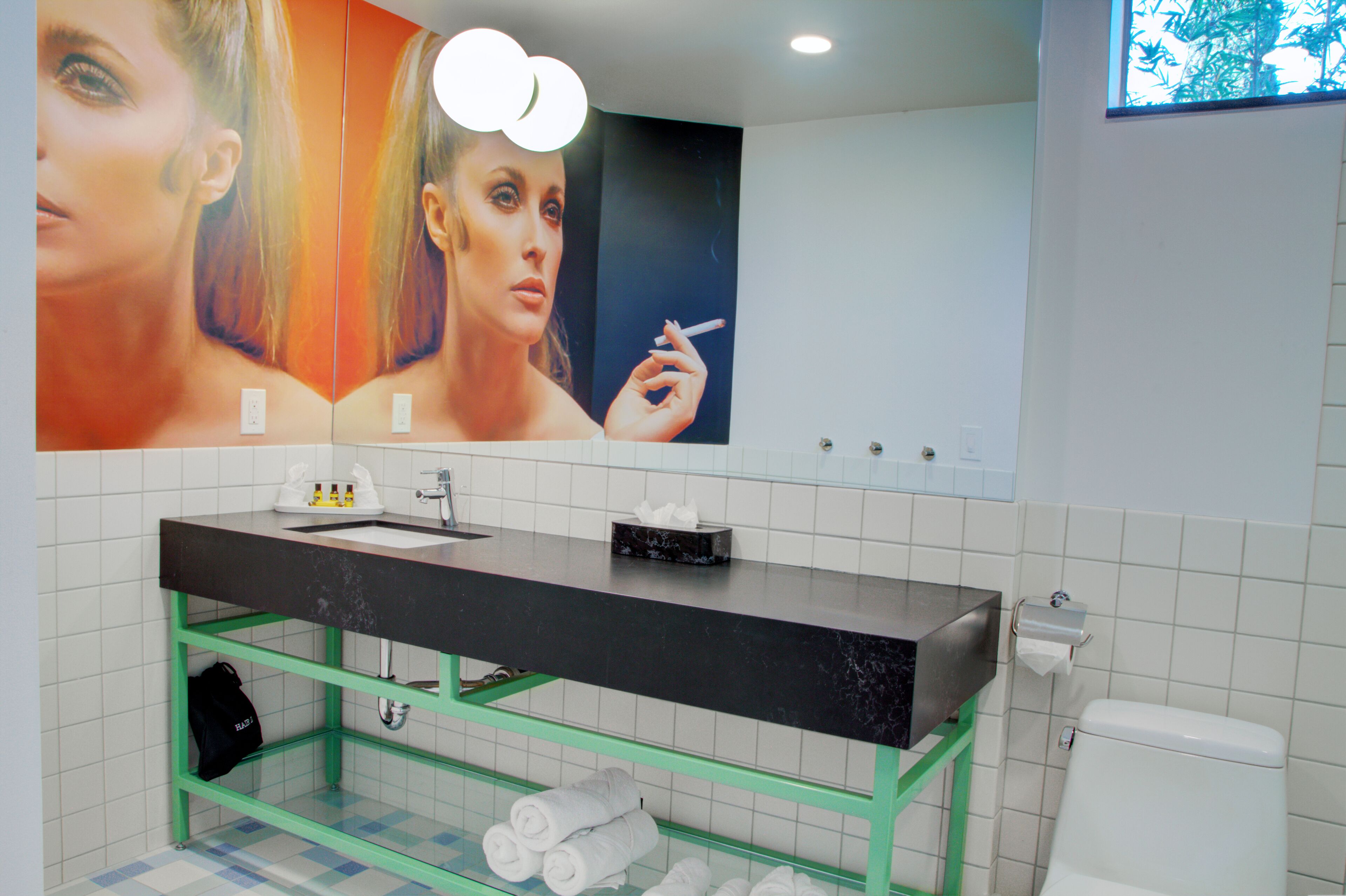 A modern bathroom with a black marble sink, green shelving, and a large mirror with a reflection of a woman holding a cigarette.