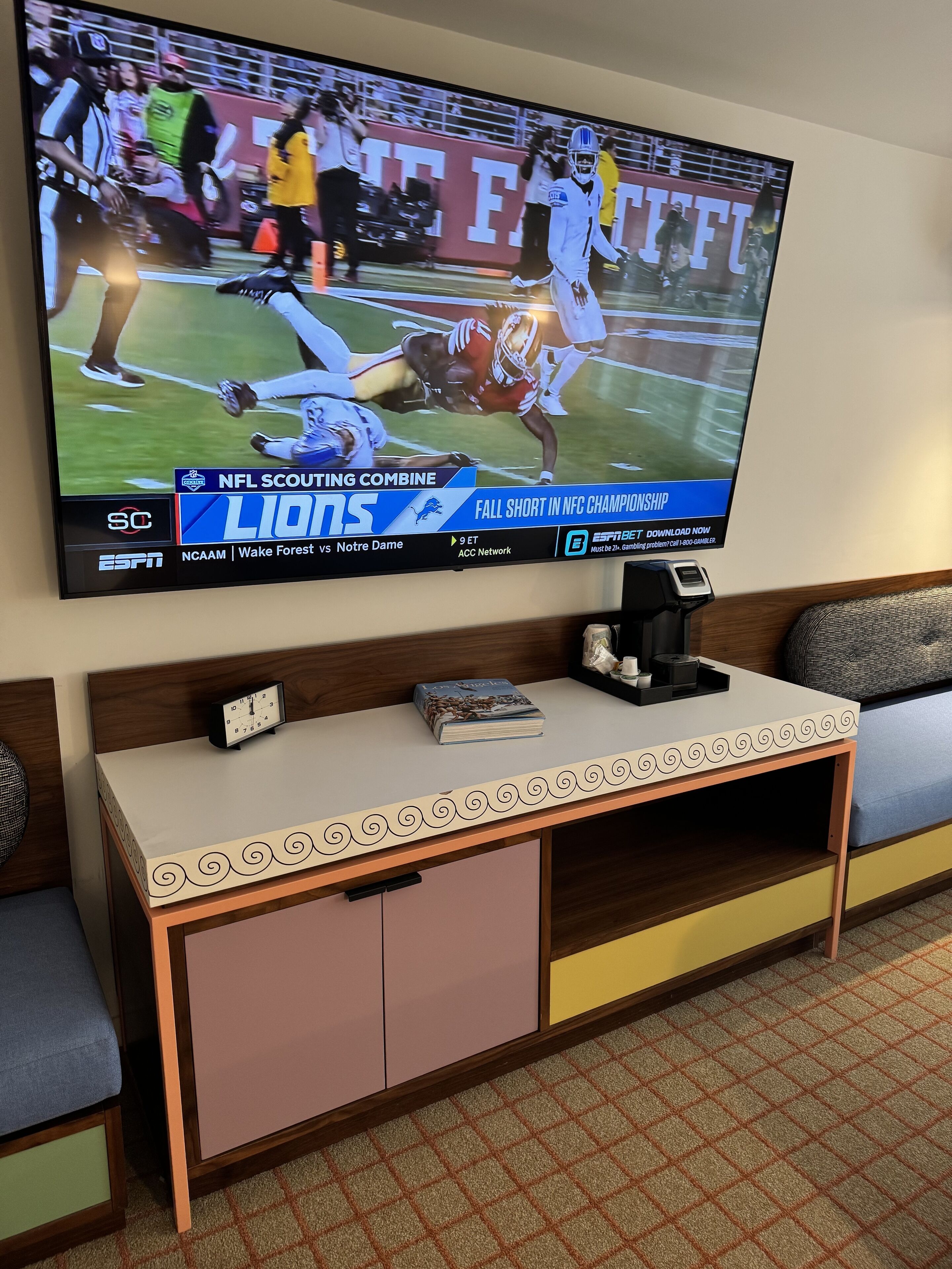 A television displaying an NFL game, a clock, and a book on a bedside table.
