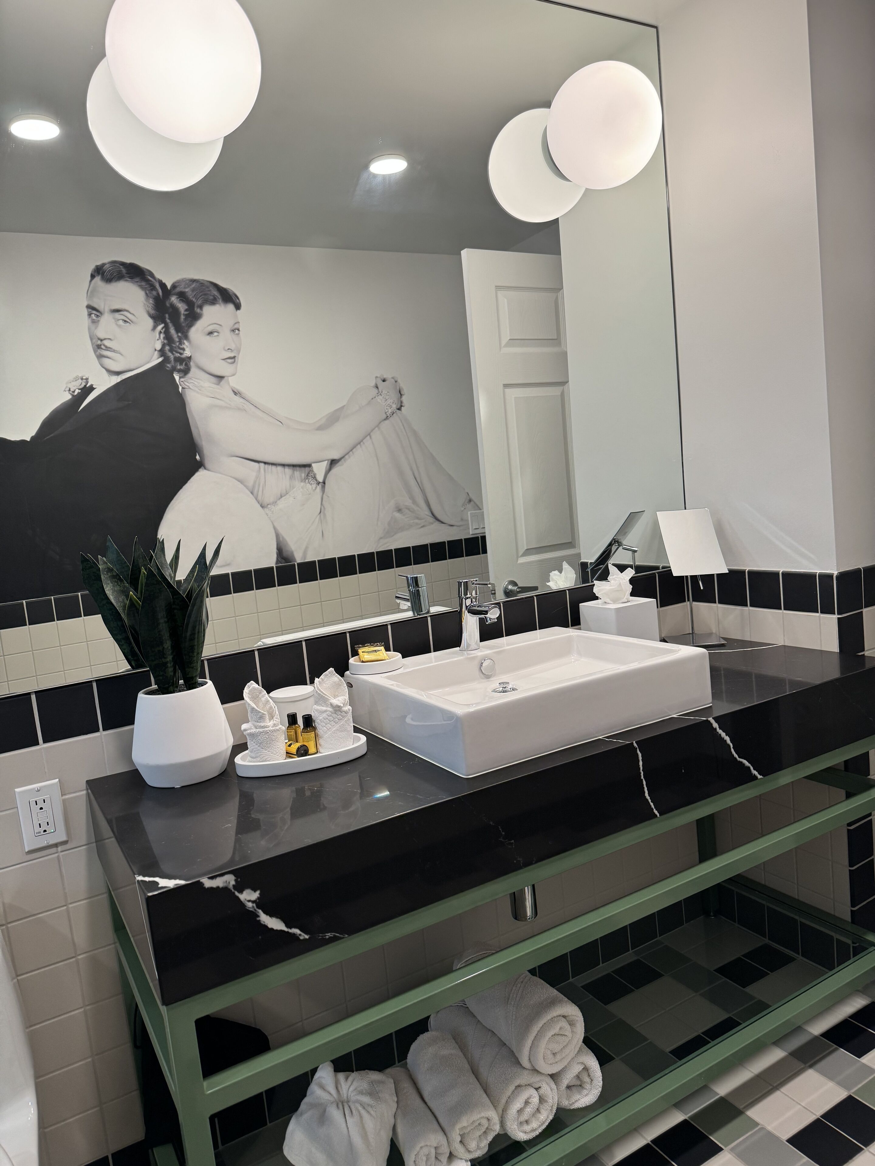 A modern bathroom with a black and white tiled backsplash, a white sink, and a glass vanity. There is a mirror above the sink, and a potted plant on the counter.