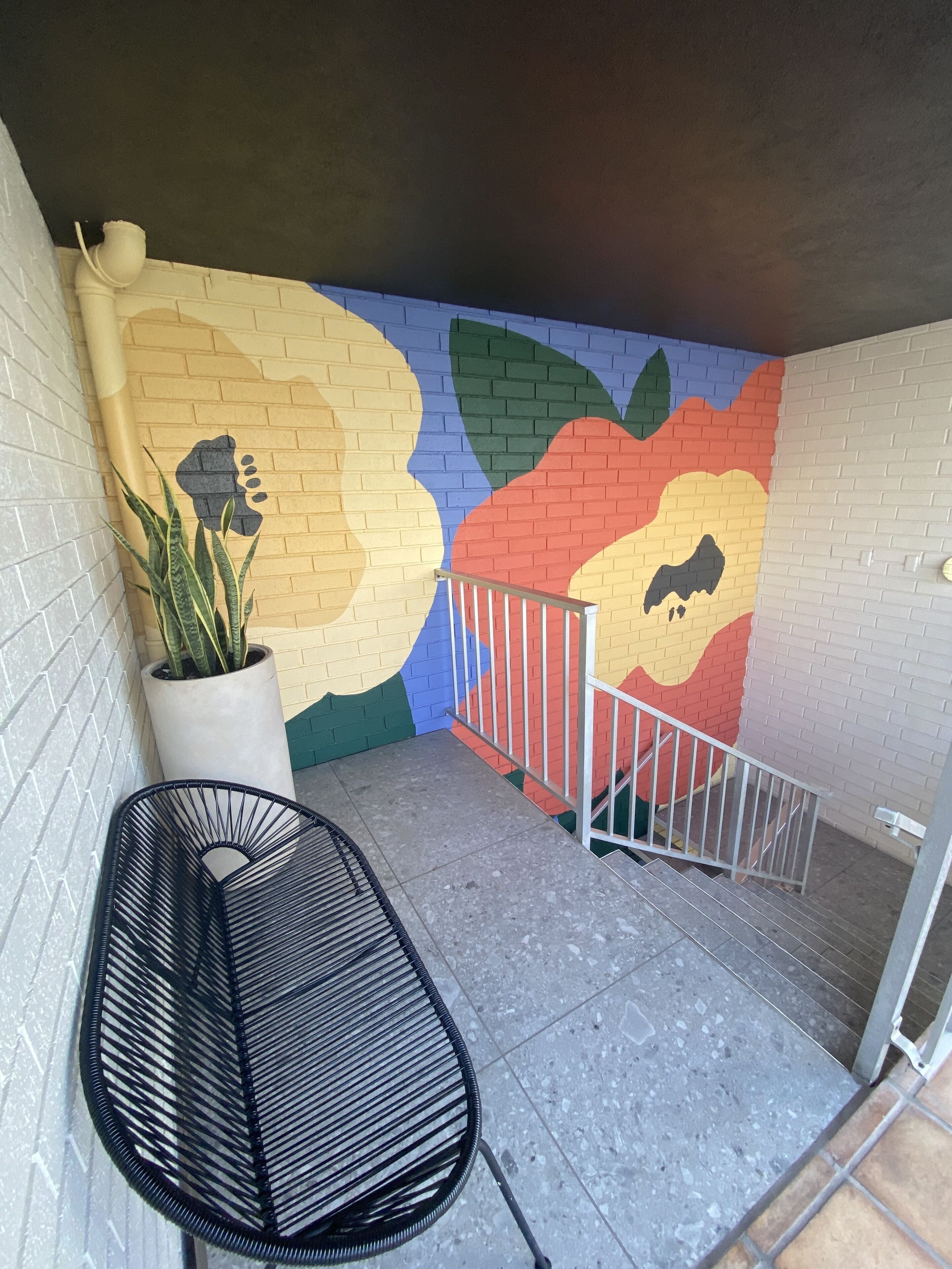 A small outdoor area with a colorful mural on the wall, a potted plant, and a black chair.