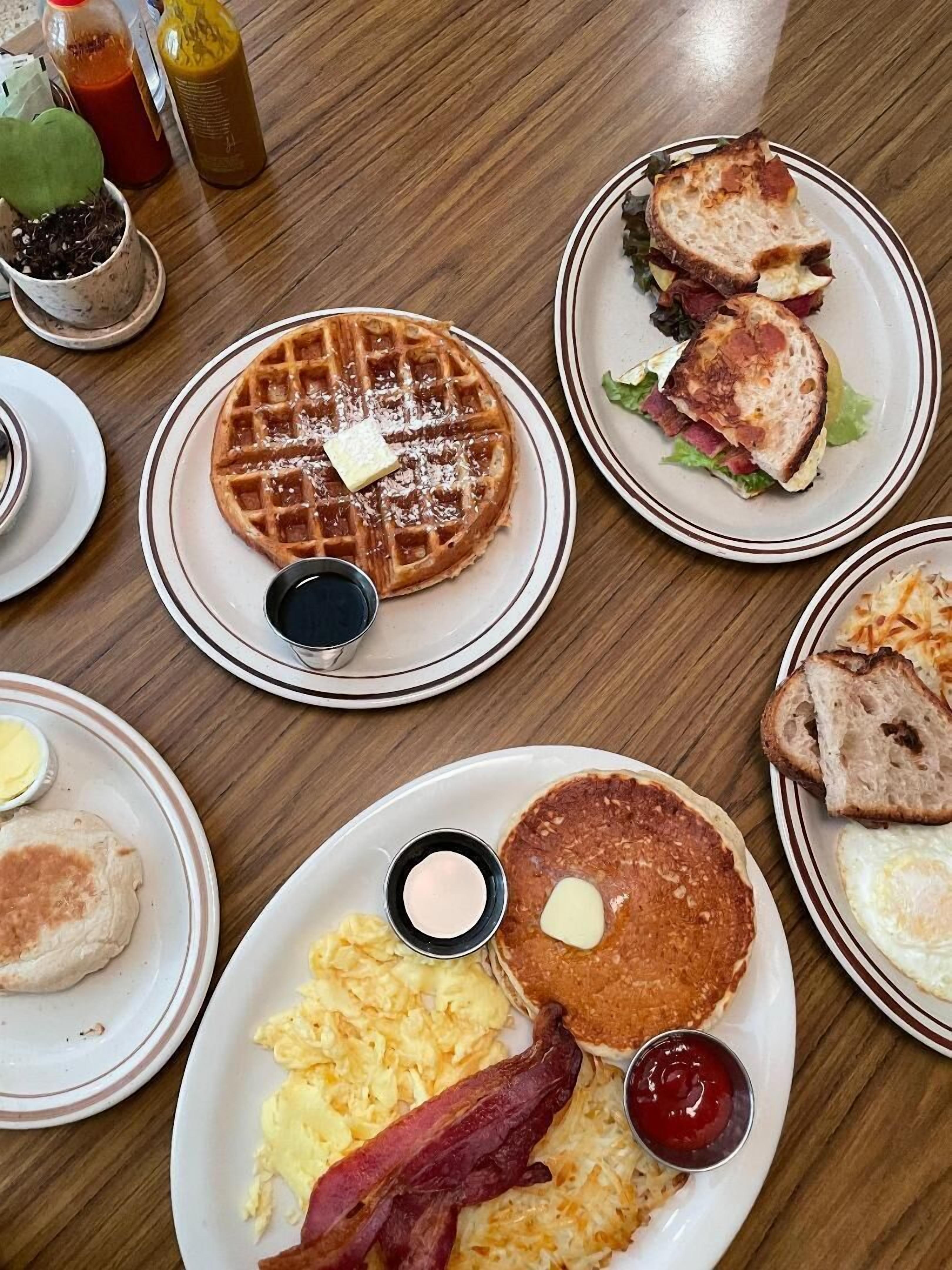 A breakfast table with waffles, sandwiches, pancakes, eggs, bacon, and condiments.