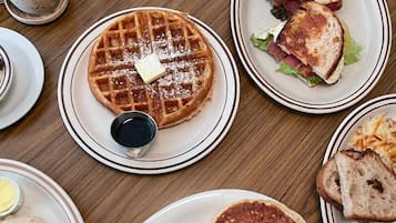 A breakfast table with waffles, sandwiches, pancakes, eggs, bacon, and condiments.