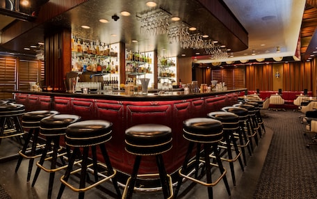 A bar area with a red cushioned bar counter, multiple stools, and a variety of bottles and glasses on shelves.