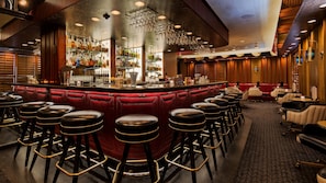 A bar area with a red cushioned bar counter, multiple stools, and a variety of bottles and glasses on shelves.