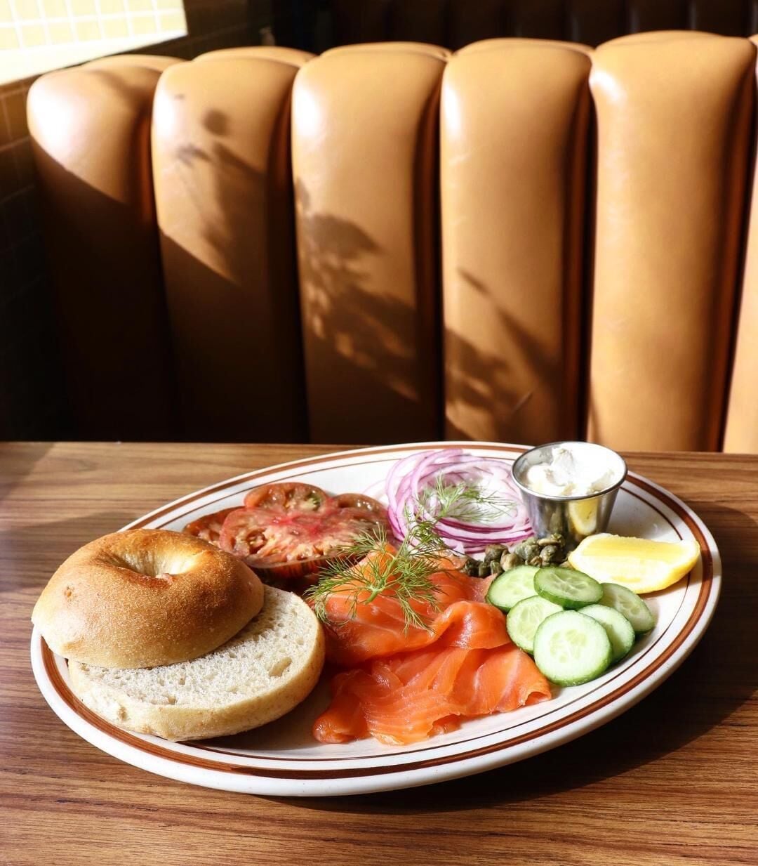 A plate with bagel, smoked salmon, cucumber slices, lemon, and a small container of cream cheese on a wooden table.