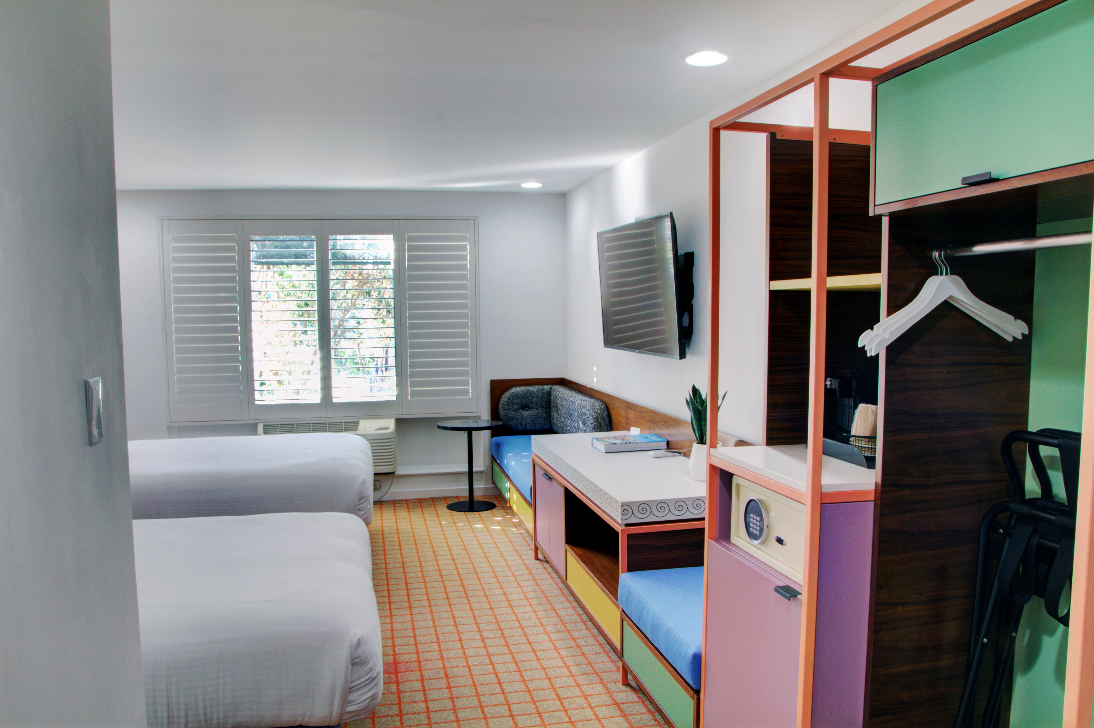 A compact room with two single beds, a small table, a bench, a TV mounted on the wall, and a wardrobe with a hanging rack.