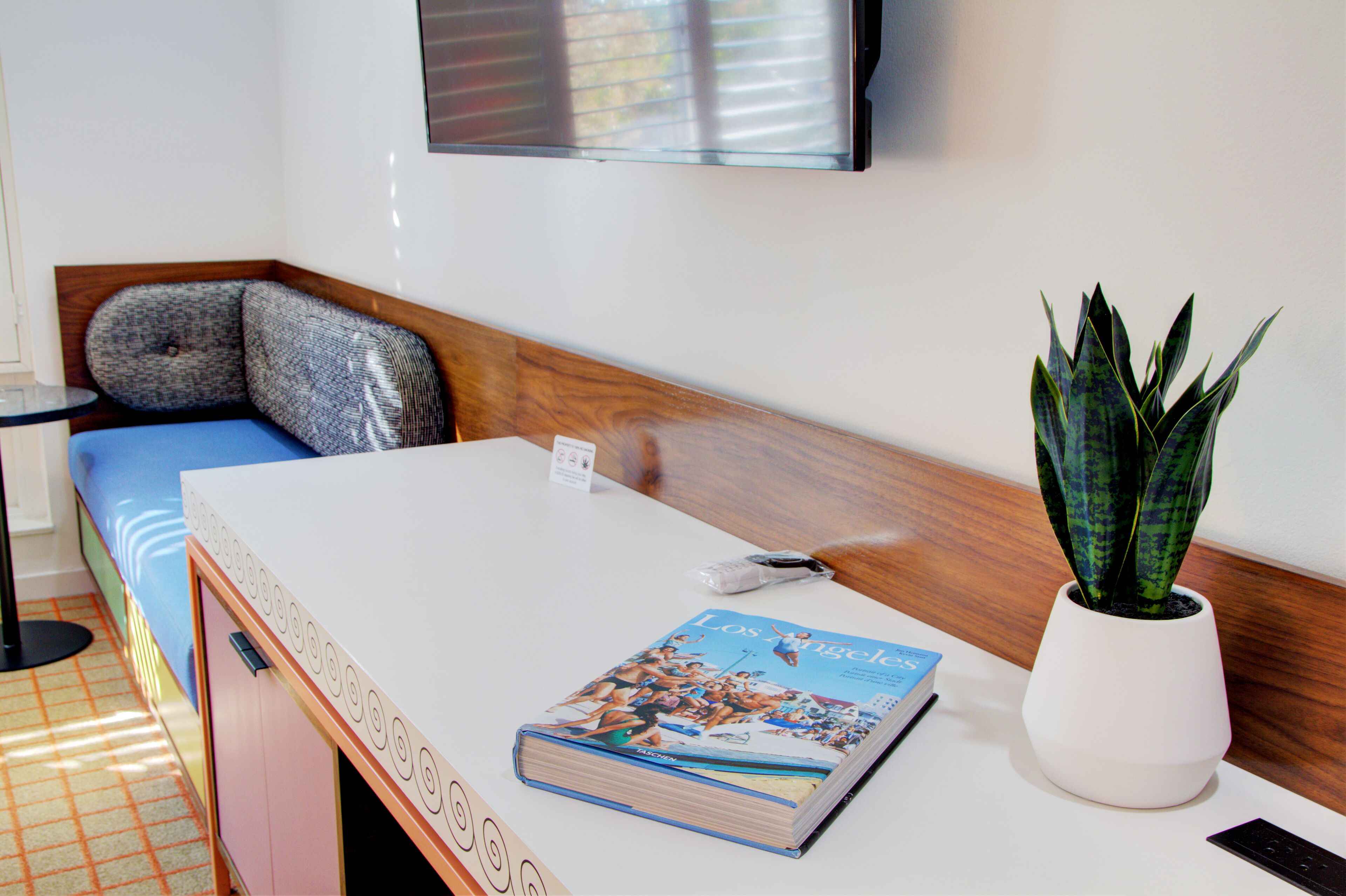 A hotel room with a bed, a small table with a magazine, a potted plant, and a wall-mounted TV.