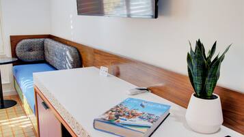 A hotel room with a bed, a small table with a magazine, a potted plant, and a wall-mounted TV.