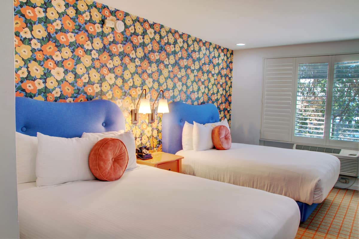 Een hotelkamer met twee bedden, een bloemen behang, een nachtkastje met lampen en een raam met zonwering.