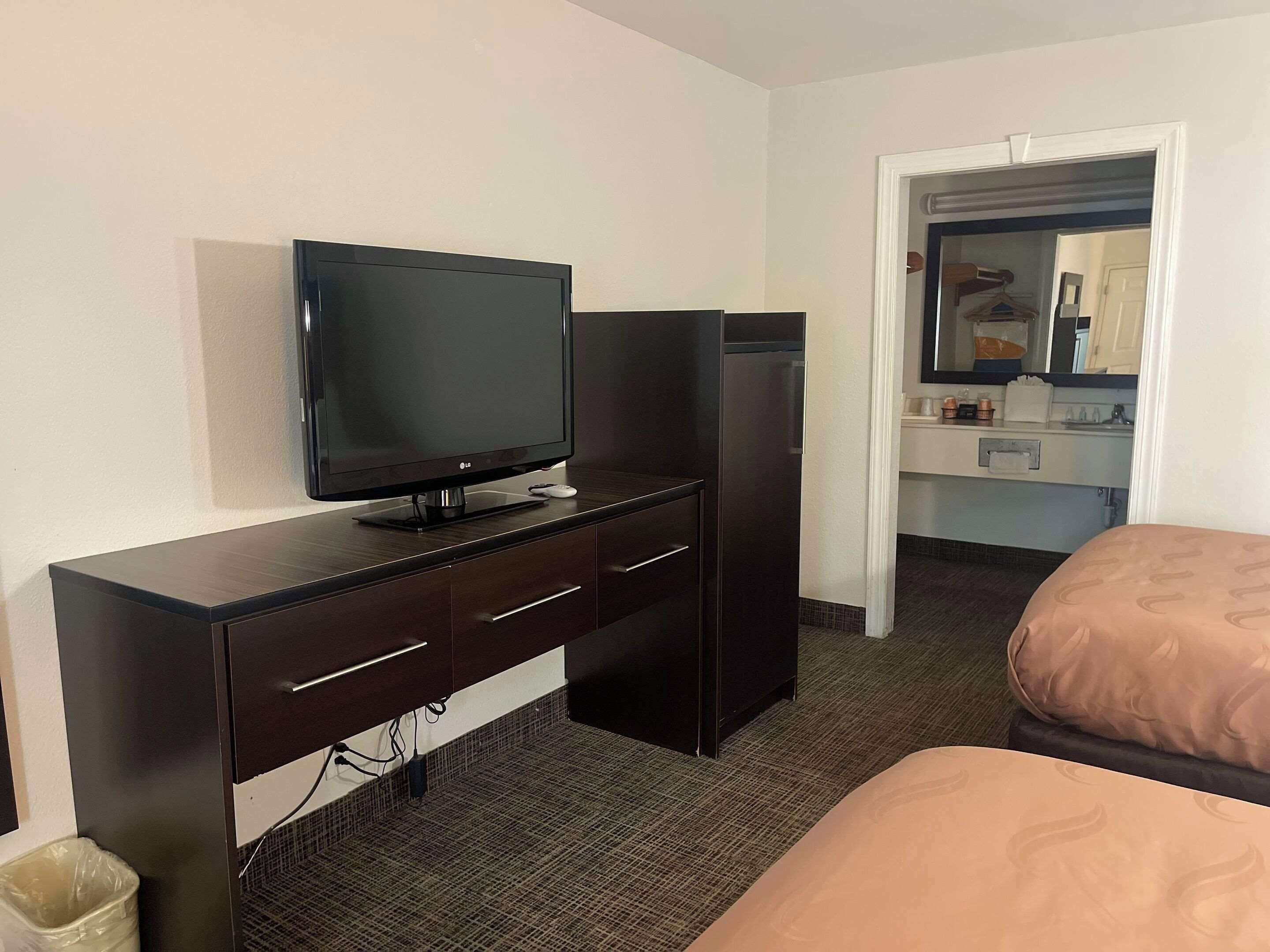 A hotel room with a flat-screen TV on a dark wooden desk, a refrigerator, a bed with a patterned cover, and a bathroom visible through an open door.