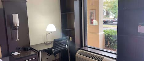 A hotel room with a desk, chair, lamp, air conditioning unit, and a window with a view of the outside.
