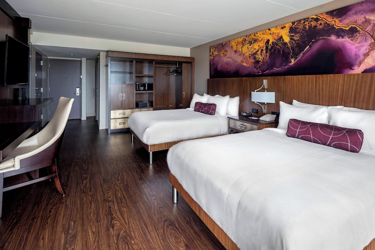 A hotel room with two beds, a desk, a chair, a TV, and a large abstract painting on the wall.