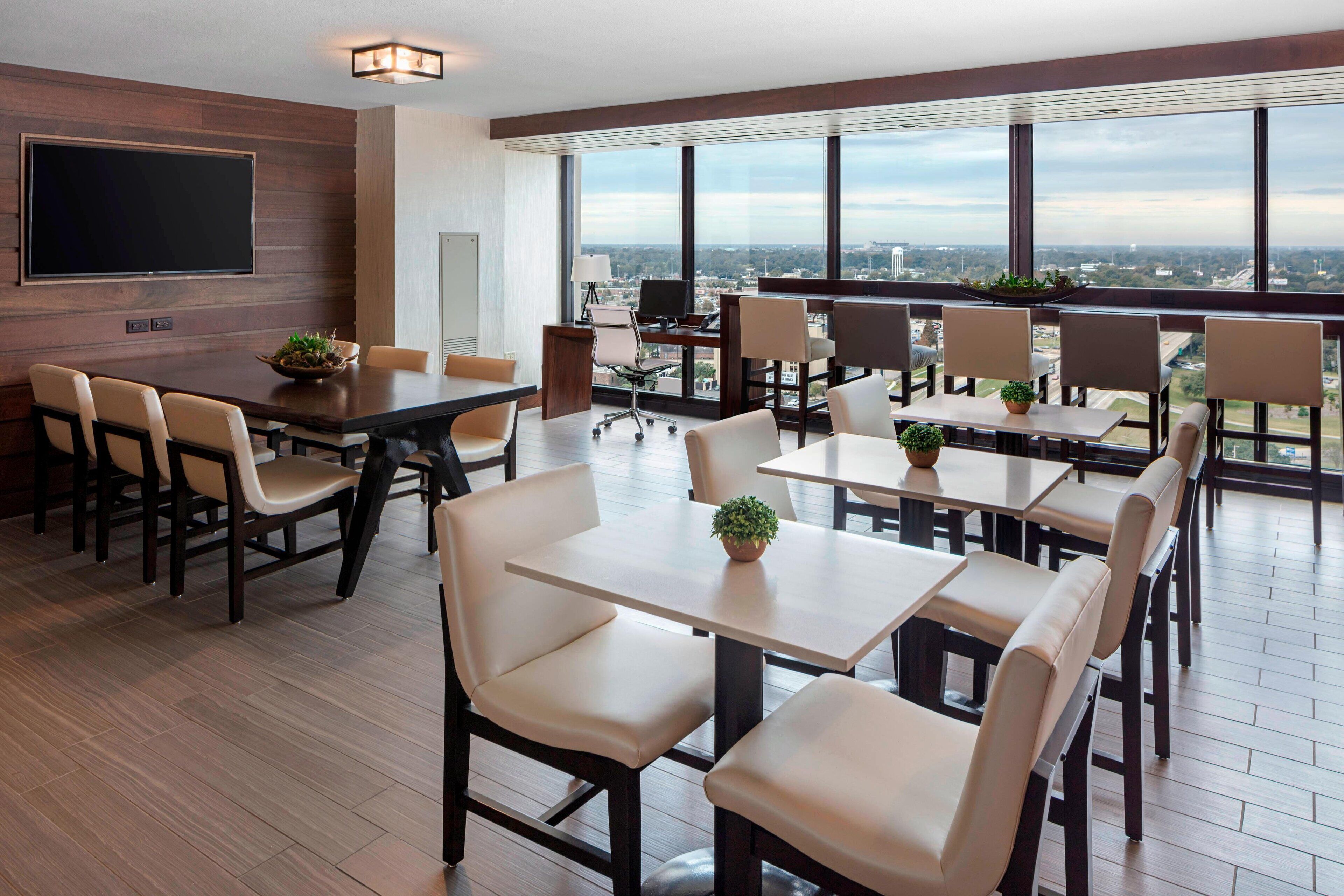 A modern conference room with a large flat-screen TV, multiple tables and chairs, and large windows offering a city view.