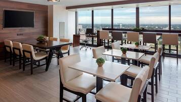 A modern conference room with a large flat-screen TV, multiple tables and chairs, and large windows offering a city view.