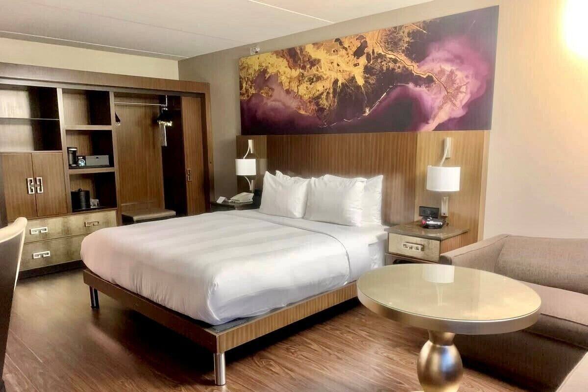 A hotel room with a bed, a desk, a chair, a sofa, a round table, and a large abstract painting on the wall.