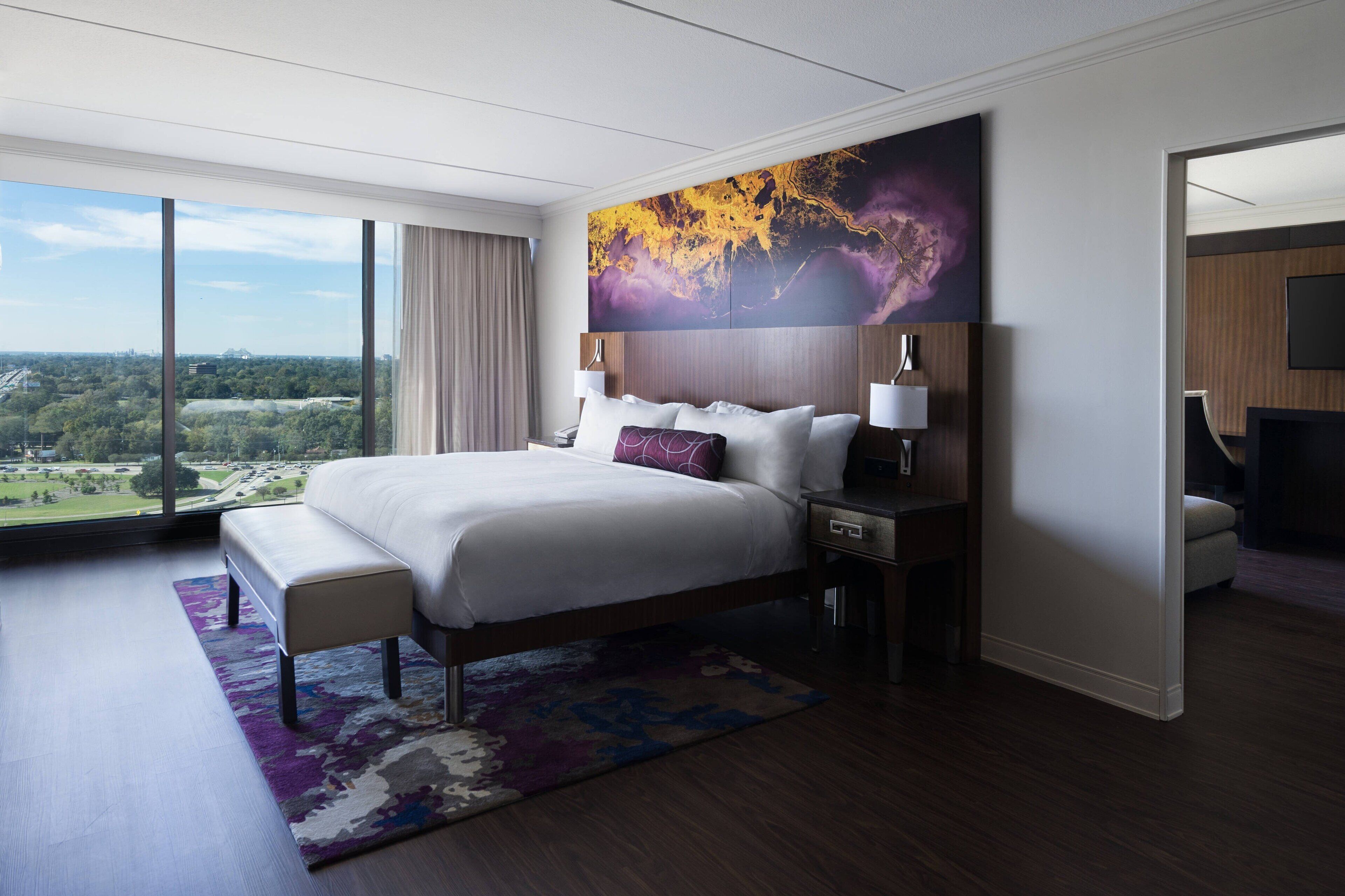 A modern hotel room with a large bed, a view of the city, and a large abstract painting on the wall.