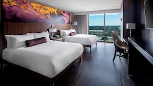 A hotel room with two beds, a desk, a chair, a TV, and a large window offering a city view.
