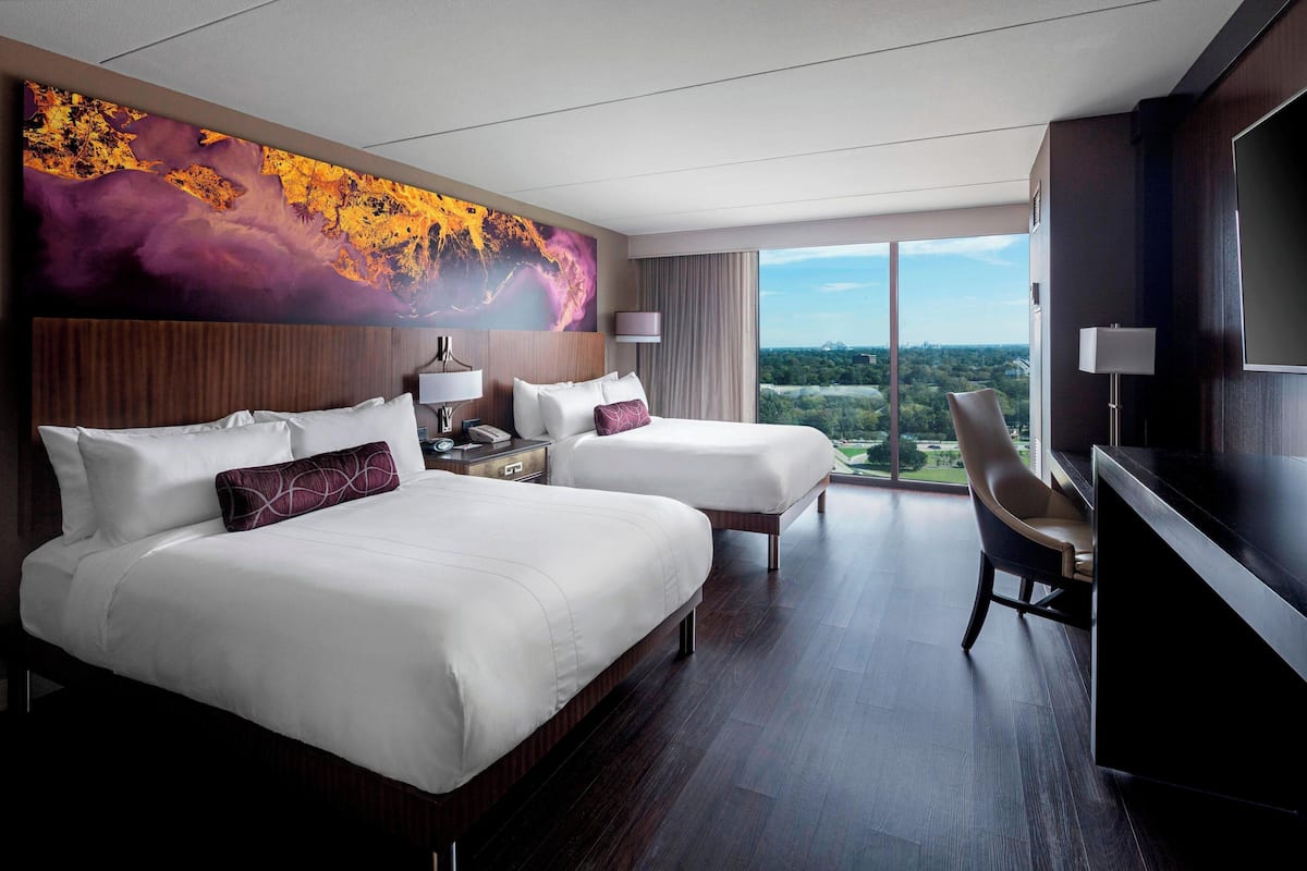 A hotel room with two beds, a desk, a chair, a TV, and a large window offering a city view.