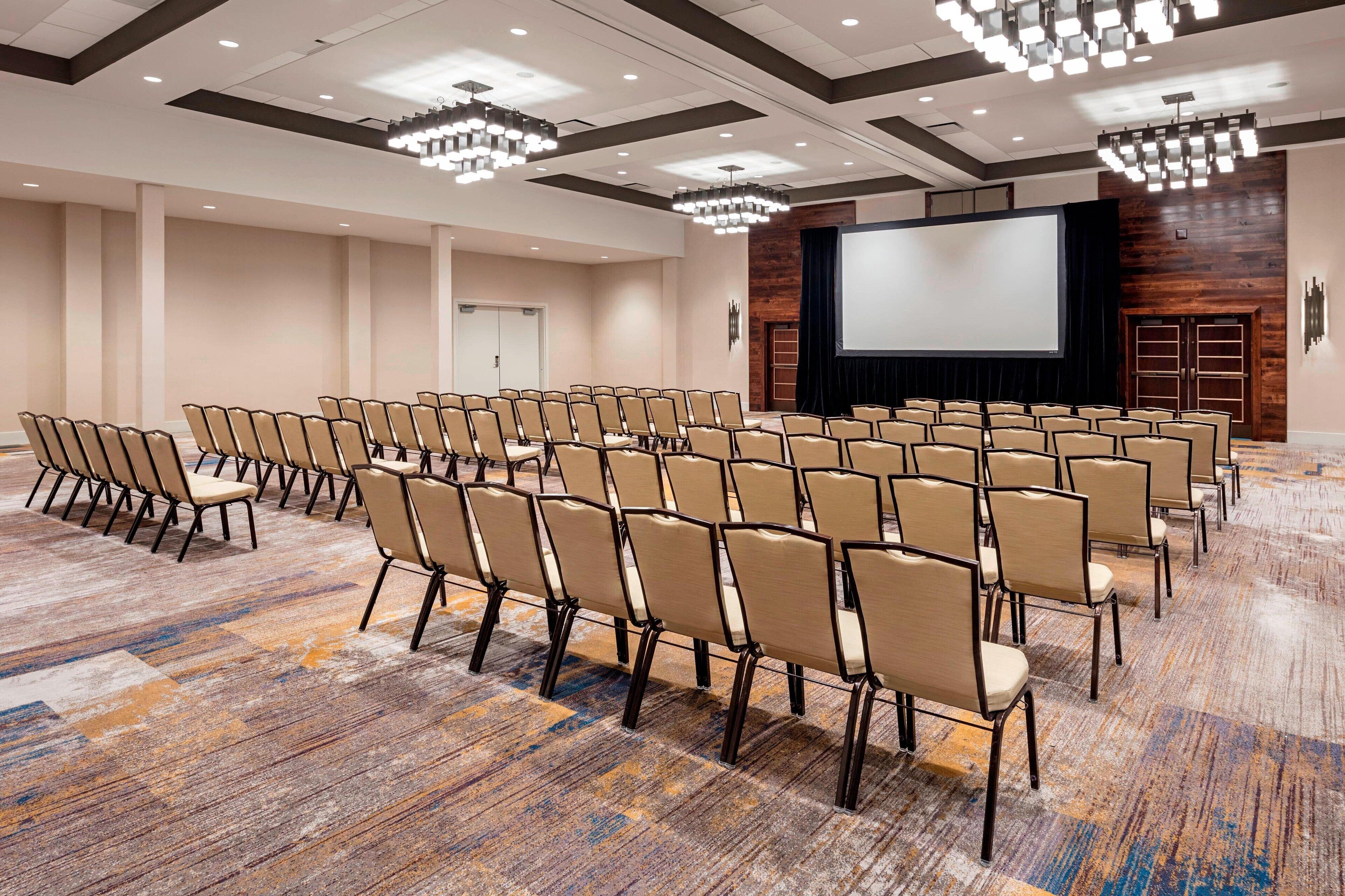 Un salón de conferencias con filas de sillas, una pantalla grande y una pared de madera decorativa.
