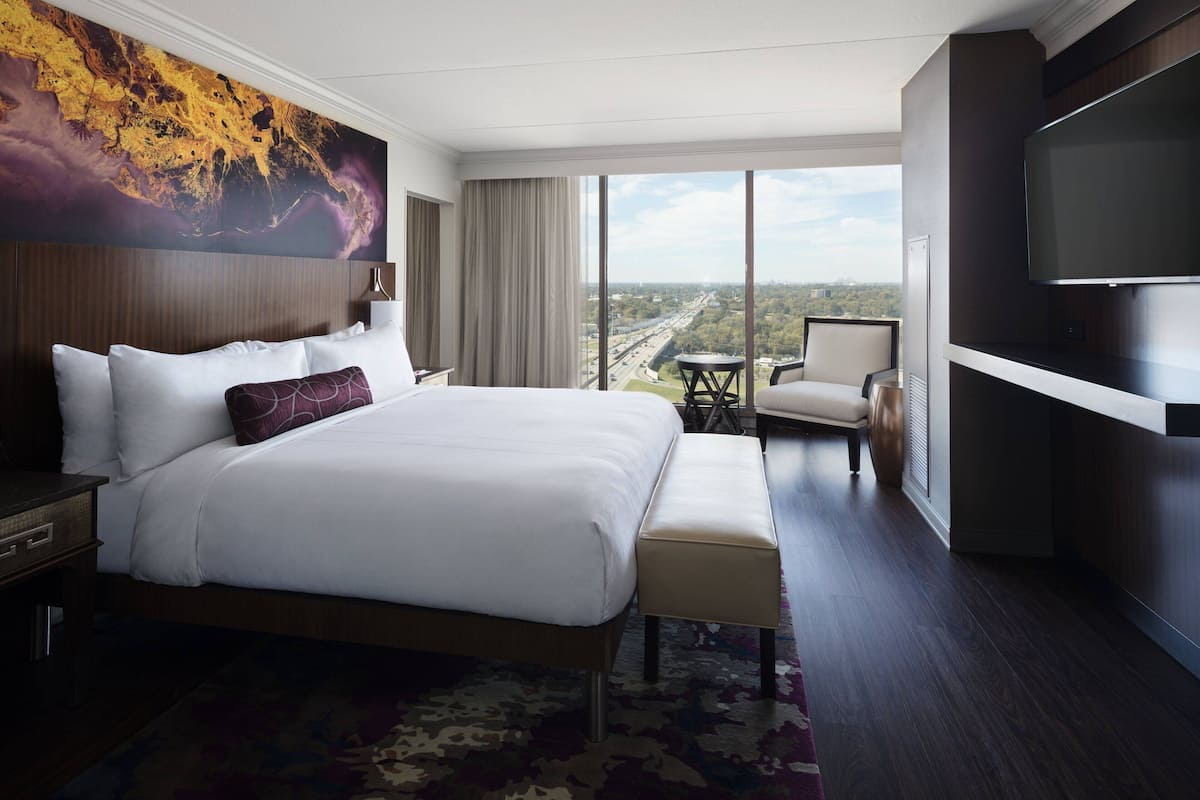 A hotel room with a large bed, a desk, a chair, a TV, and a view of the city.