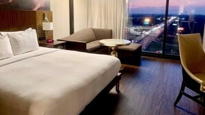 A hotel room with a large bed, a desk, a chair, and a view of a cityscape at dusk.